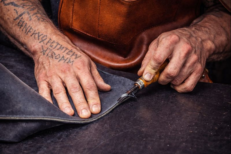 Handwerkliche Lederverarbeitung: muskulöse Hände mit Runen-Tattoo schneiden Echtleder aus Rindsleder mit traditionellem Werkzeug